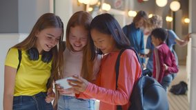 Three multiethnic school girlfriends using smartphone in corridor during break. Diverse schoolgirls surfing internet on mobile phone together in hallway after classes - Powered by Shutterstock - Get 15% off with code: PIKWIZARD15