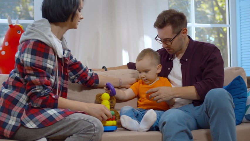 Happy mother, father and baby son playing with pyramid toy at home. Portrait of cheerful young parents and toddler boy sitting on couch at home playing colorful toys