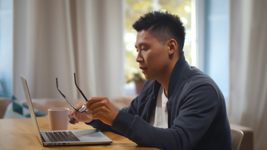 Tired asian man putting off glasses and rubbing eyes working on laptop in home office. Side view of overworked freelancer using laptop and drinking coffee at table in living room | Shutterstock HD Video #1060591585