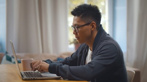 Handsome young asian man wearing casual clothes sitting down at table with cup of coffee using laptop computer at home. Korean freelancer in glasses working at home office