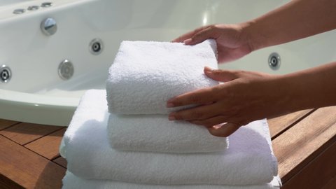 Woman puts one clean soft white towel on stack of folded several towels near empty whirlpool bath.