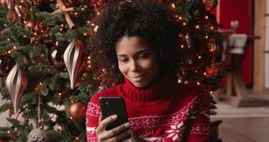 Beautiful african woman sit near decorated with twinkled lights glowing tree using smartphone chatting with friend read New Year congrats laughs feels happy. Christmas sale e-commerce user concept