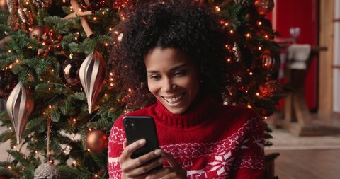 Beautiful african woman sit near decorated with twinkled lights glowing tree using smartphone chatting with friend read New Year congrats laughs feels happy. Christmas sale e-commerce user concept Video de stock
