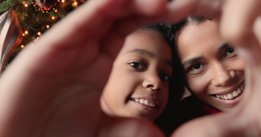 Close up African mom and daughter faces view through heart shape joined fingers, vloggers share love congratulate subscribers Merry Christmas. New Year celebration, happy holidays, best wishes concept | Shutterstock HD Video #1060593400
