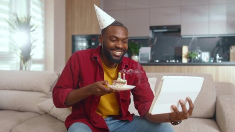 Excited African american businessman video chatting on webcam tablet conferencing with family blowing candles on birthday cake celebrating holiday at home alone. Internet Relationships. Communication