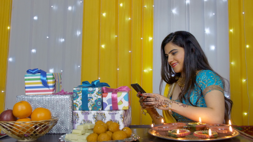 Attractive Indian girl using her smartphone while wearing a traditional lehenga. Beautiful young female replying to the festival messages while sitting around Diwali gifts, oil lamps, fruits, and s...