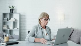 Middle Aged Woman With Blond Hair Sitting At Table In The Apartment And Talking To Someone Via Video Link. She Has Headphones In Her Ears. Woman Picks Up The Laptop And Turns It In Different
