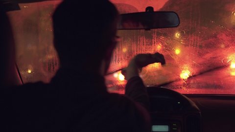 Driver cleaning the car glass during a heavy rain. Red lights of the traffic jam from the interior of a vehicle. Cinematic storm inside a car.