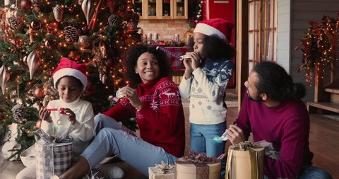 Happy African family with kids gathered together in modern living room near glowing decorated Christmas tree and heap of gift boxes blowing party whistles celebrating 2021 New Year. Holidays concept