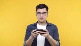 A man put on glasses and denim jacket white t-shirt is playing game on smartphone using modern technology - apps, social networks feel headache isolated on yellow background.