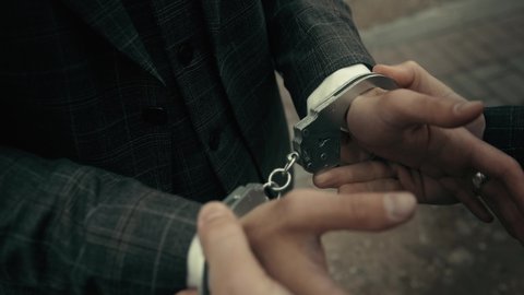 Arrest of the offender without resistance. Policeman puts the handcuffs on the hands of criminal, hands closeup. Detention of the accused business person.