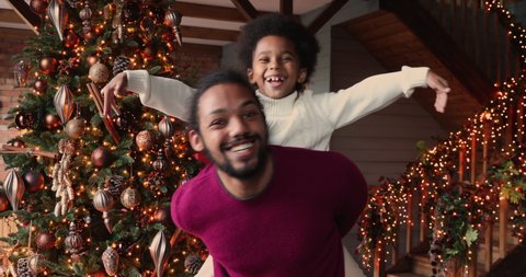 Active african father carrying riding on back little son, kid boy stretched arms like plane wings, family fool around on Christmas Eve at modern cozy decorated home. Happy holidays, have fun concept