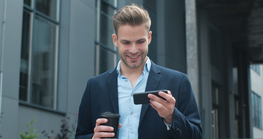 Handsome Caucasian Businessman is laughing while standing outdoor with Smartphone. Young Male is watching some Comedy Films, using Smartphone, browsing funny Apps. Business People. Phones. App.