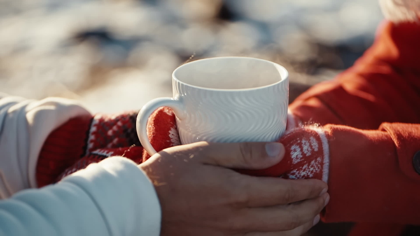 Close up hands of couple holding cups with hot tea at winter days. Coffee in the hands of a girl and a guy at xmas date. Freezing weather and warm hugs. Love concept. | Shutterstock HD Video #1060604356