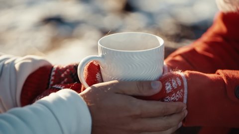 Close up hands of couple holding cups with hot tea at winter days. Coffee in the hands of a girl and a guy at xmas date. Freezing weather and warm hugs. Love concept.