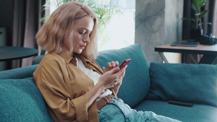 Attractive blond young woman sitting on a blue sofa using smartphone in modern room. Scrolling content bloggs. Communication, message portrait internet. Slow motion Royalty-Free Stock Footage #1060604986
