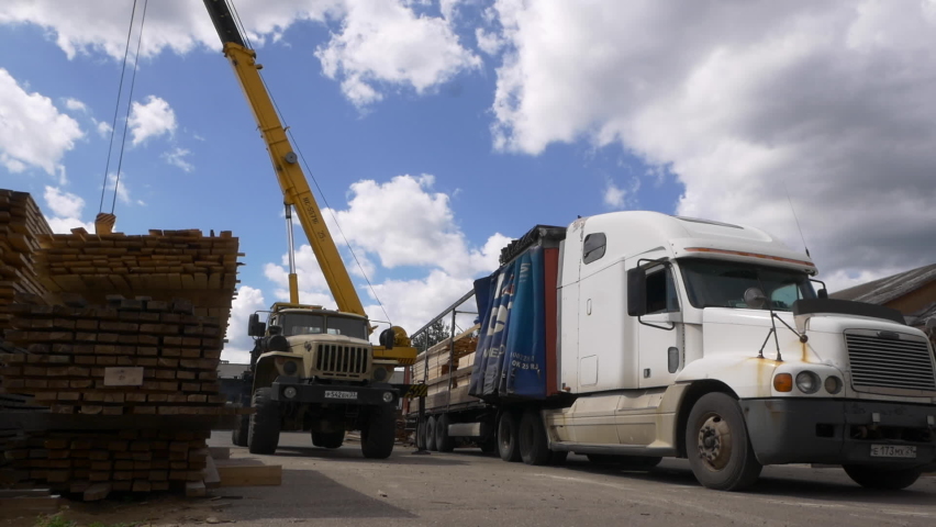 Truck crane loads wooden planks into huge lorry workbody in production factory yard under cloudy sky on autumn day Royalty-Free Stock Footage #1060607707