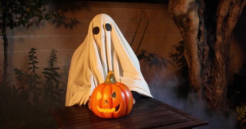Person disguised as a ghost sitting at a table with a Halloween pumpkin