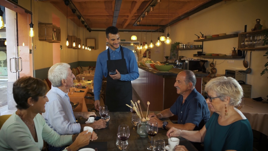 Smiling senior man sitting on table with friends paying with smart phone using NFC technology. Afro american waiter standing and holding a card terminal in a restaurant. Contactless payment.