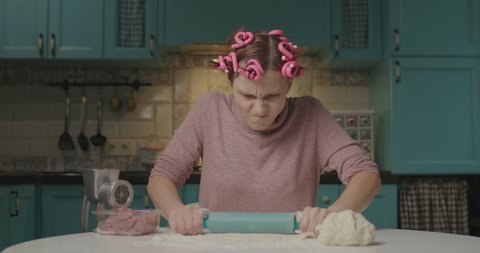 Young angry adult woman rolling out the dough with a rolling pin sitting in kitchen. Housewife with hair curler tired of cooking at home alone. Woman looking angry at camera.