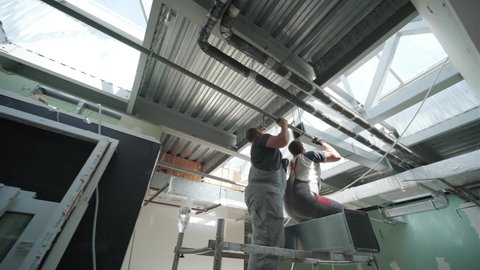 HVAC Installers On Scaffold Assembling Air Ventilation Shaft In Silver Material Hanging From Ceiling Inside New Building.