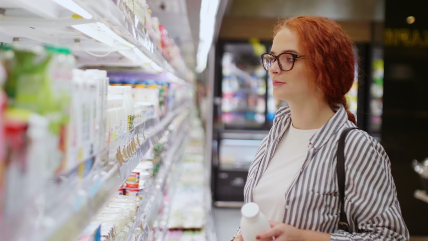 Pretty redhead girl in eyeglasses dressed casual wear choosing milk and smiling on camera in modern grocery store. Supermarket concept Royalty-Free Stock Footage #1060614268