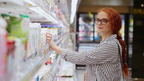 Pretty redhead girl in eyeglasses dressed casual wear choosing milk and smiling on camera in modern grocery store. Supermarket concept