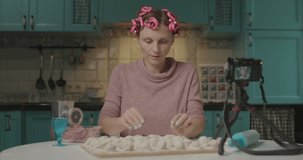 Female food blogger cooking dumplings and recording video on camera. Smiling young woman with hair curler showing the result of her cooking to social media followers.