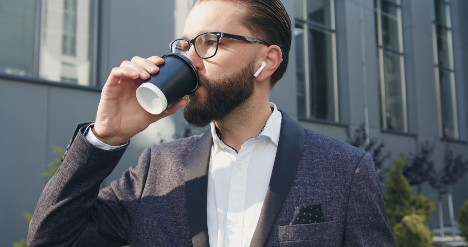 Close up of good-looking stylish thoughtful bearded man in glasses which drinking coffee near modern urban building while going to his job,4k | Shutterstock HD Video #1060619140