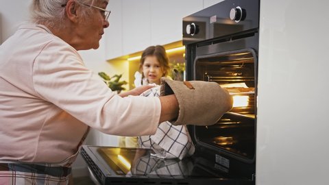 Gray-haired grandmother is taking out hot cookies from the oven, surprised granddaughter is looking at it. Baking homemade biscuits at modern kitchen. Happy family, relationship. Close up, slow motion