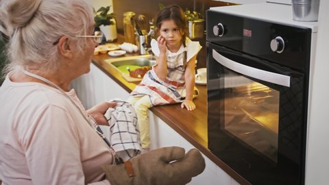 Gray-haired granny is opening the oven and taking out hot cookies, surprised grandchild is looking at it. Baking homemade biscuits at modern kitchen. Happy family, relationship. Close up