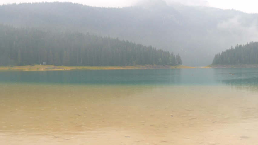 Black lake in Montenegro during the rain. Raindrops fall on the water surface. The mesmerizing beauty of the pristine nature of the lake, mountains and forest.