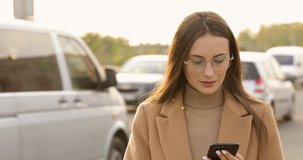 Portrait of stylish young lady in eyeglasses using smartphone while standing near road with lots of moving cars. Pretty woman in beige coat using modern gadget outdoors.