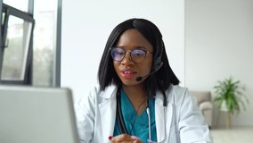 Professional african american female doctor in white medical coat and headset making conference call on laptop computer, consulting distance patient online in video chat