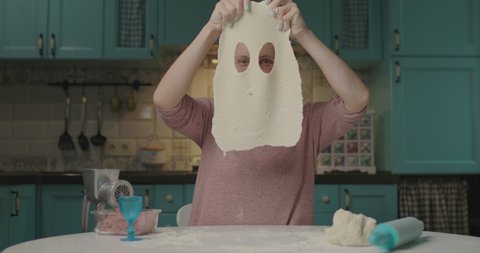 Young adult woman making holes in dough sitting in kitchen. Smiling housewife with hair curler cooking at home alone. Dough funny mask. 