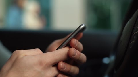 Extreme close-up hands of unrecognizable man typing online message using mobile phone while sitting at car. Closeup view of businessman using phone in auto. Random passers-by walk on the street.