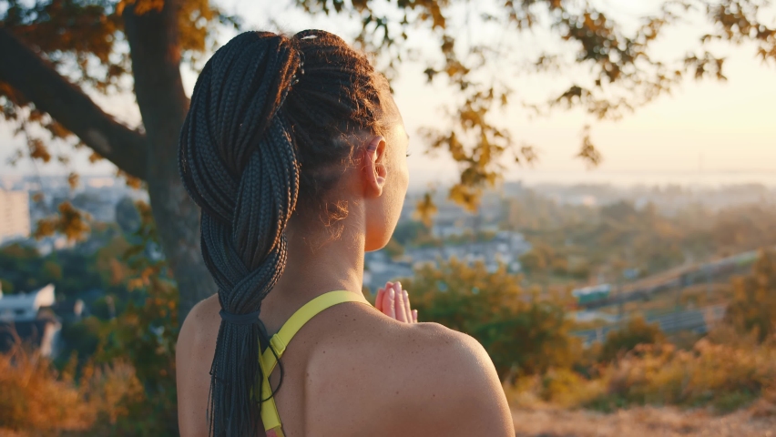 Slim beautiful young woman with black braids holding hands together performing yoga pose meditating on hill landscape in summer sunset. Stylish girl. Active lifestyle. Yoga training. Royalty-Free Stock Footage #1060629535