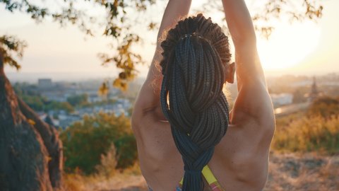 Slim beautiful young woman with black braids holding hands together performing yoga pose meditating on hill landscape in summer sunset. Stylish girl. Active lifestyle. Yoga training.