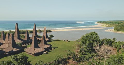 Aerial view of traditional village with ornately houses roof on sand beach sea shore with tropic trees and plants. Green grass valley at sandy ocean coast Sumba Island, Indonesia, Asia at drone shot