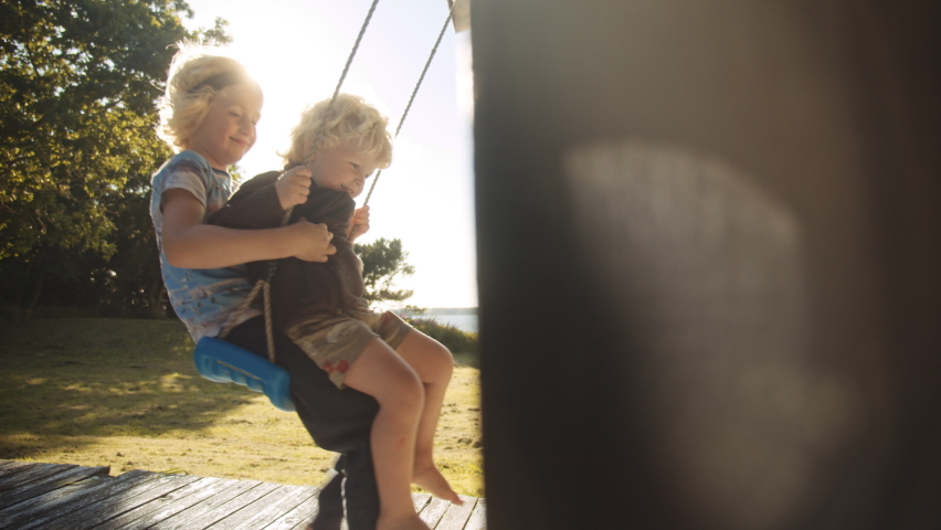 Slow Motion Wide Handheld Shot Of Two Young Brothers Sitting On Swing On Sunny Summer