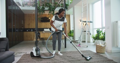 Happy cheerful young beautiful african american woman using modern vacuum cleaning carpet floor in a bright cozy living room at home. Concept of housekeeping, cleaning house, lifestyle.