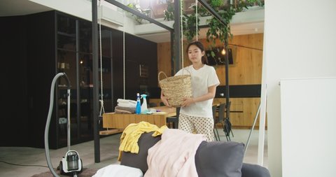 Close up of happy cheerful young beautiful asian woman with basket picking dirty laundry in cozy living room at home. Concept of housekeeping, housework, cleaning house, lifestyle.