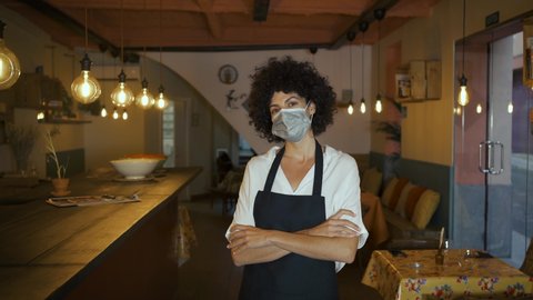 Confident female owner standing in the dining room of her restaurant, crossing arms and looking at camera. Waitress wearing apron and surgical face mask. Small business, people and crisis concept.