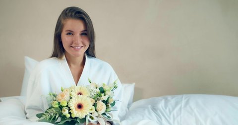 Happy young lady in bathrobe holds fresh flowers bouquet sitting on large comfortable bed in hotel room in sunny morning