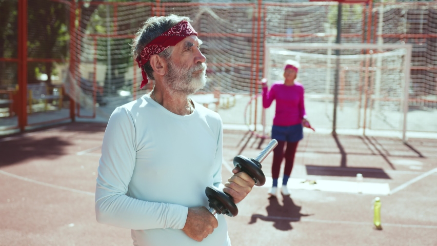 Serious elderly athlete posing with heavy dumbbells on training session outdoor. His fit wife jumping on rope doing active physical exercises. Sports team. Concept of family.