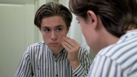 Young guy crushes pimples in the bathroom. Awkward age. Problems with the skin. Difficulties of teenagers. Dermatology