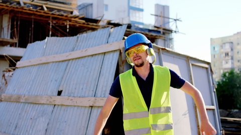 Man construction worker with a safety helmet and goggles listening music from the headphones at the construction site while have a break time in front of the camera