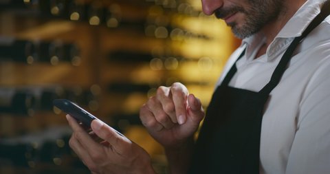 Modern farmer or winemaker is using winery online commerce applications on a smartphone for checking customer service and selling orders summary of his wine production in a wine cellar.