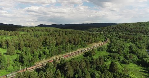 Freight long train carries with coal carriages an electric locomotive by dangerous part of two-sided Trans Siberian railways in forest mountains / Aerial drone wide view at summer sunny day