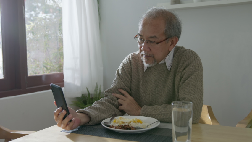 Medium shot : Retired gray haired grandpa asian man sit alone using mobile phone video call at table desk at window boring stay home self isolation quarantine feeling depress in problem mental health.